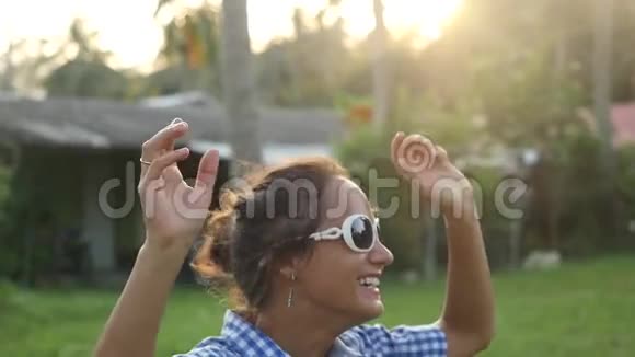 快乐时尚的年轻女子戴着太阳镜在阳光下跳舞在热带棕榈树之间在丛林中唱歌慢动作视频的预览图