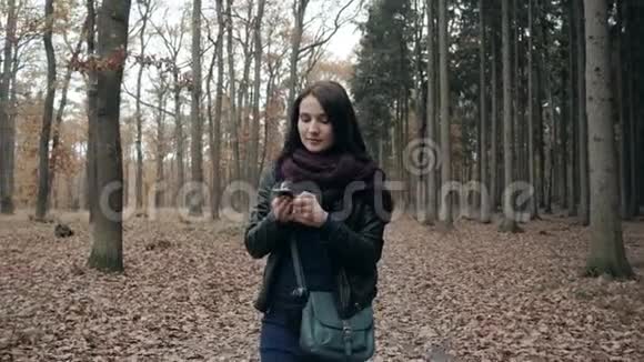 美丽的年轻的布鲁内特女士使用智能手机在秋季公园秋天在森林里散步的女孩的生活方式概念视频的预览图