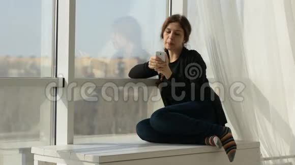 快乐的年轻女子坐在窗前在社交网络上发短信视频的预览图