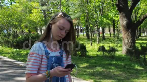 一个女孩带着耳机在公园里散步一个十几岁的女孩在户外听音乐一个少年在阳光明媚的阳光下带着手机走路视频的预览图