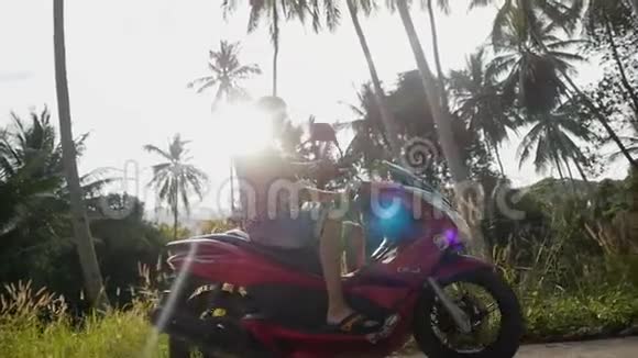 戴墨镜的青年旅行者骑着摩托车在热带阳光明媚的道路上穿过太阳美丽的镜头耀斑视频的预览图