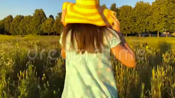 美丽的女孩戴着黄色的帽子在日落的天空上的绿色麦田上奔跑自由概念日落时的麦田视频的预览图