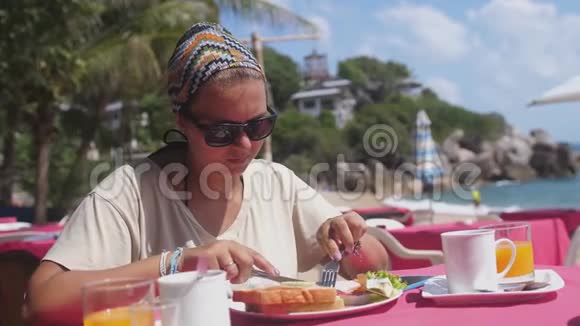 戴墨镜的悲伤女人在海边的海滩餐厅用黄油涂面包临近美味的美式早餐视频的预览图