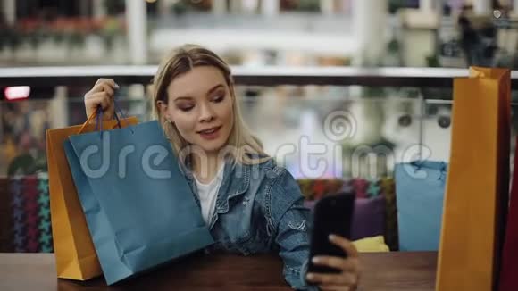 一个漂亮的金发女孩拿着购物袋坐在咖啡馆里用手机自拍视频的预览图