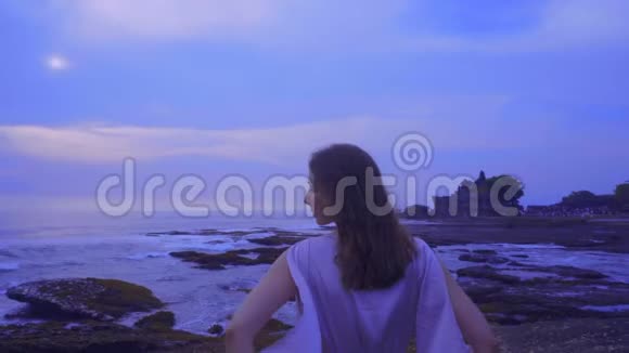美丽的年轻女性穿着一件长裙子站在巴厘岛海岸的岩石上日落时分美丽的景色尽收眼底视频的预览图