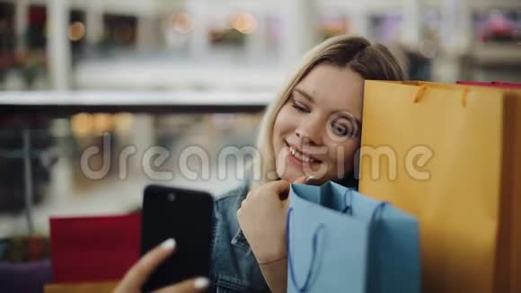 一个漂亮的金发女孩拿着购物袋坐在咖啡馆里用手机自拍视频的预览图
