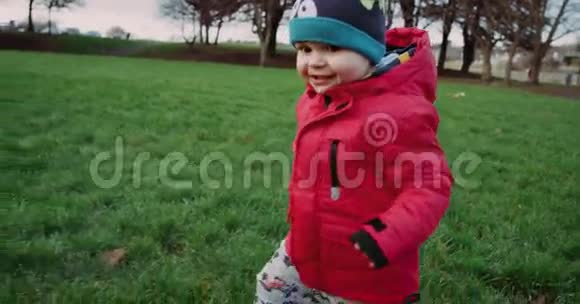 穿着红色夹克的快乐孩子在草地上的公园里奔跑和微笑4k录像慢动作视频的预览图