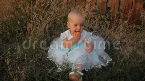 一个小女孩坐在高高的草地上穿着白色的连衣裙吃着杏子微笑着摆动着她的腿日落视频的预览图