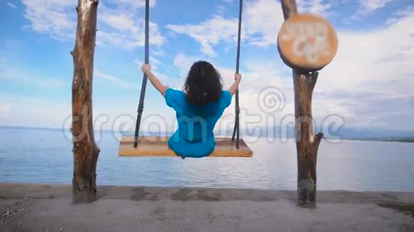 巴厘岛的海洋海滩女孩荡秋千青绿色的海洋晴空万里穿蓝色连衣裙的女孩在秋千上从这个地方视频的预览图