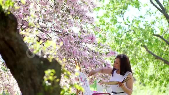 一位美丽的女画家穿着白色的衣服画家在盛开的春天苹果园里画了一幅花她视频的预览图