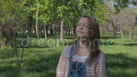 一个女孩带着耳机在公园里散步一个十几岁的女孩在户外听音乐一个少年在阳光明媚的阳光下带着手机走路视频的预览图