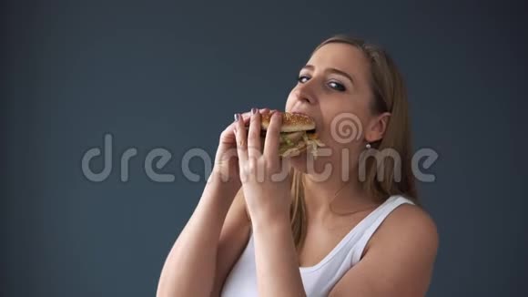 超重的女人吃汉堡包看着灰色背景的相机有害食物的概念过量视频的预览图