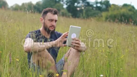 一个留胡子的年轻人坐在乡间别墅附近的草地上用平板电脑视频的预览图