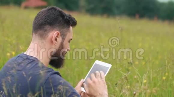 一个留胡子的年轻人坐在乡间别墅附近的草地上用平板电脑视频的预览图