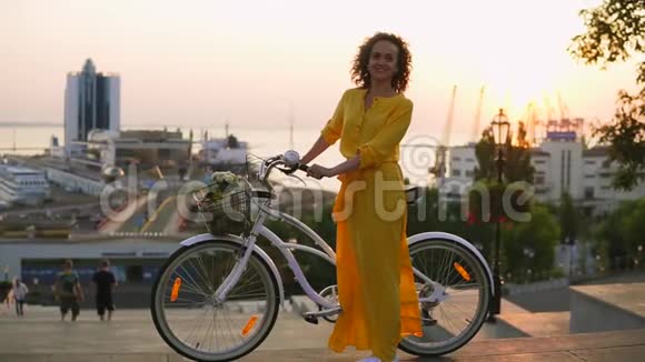 年轻的黑发女人穿着黄色长裙站在她的城市自行车旁手里拿着篮子里的花视频的预览图