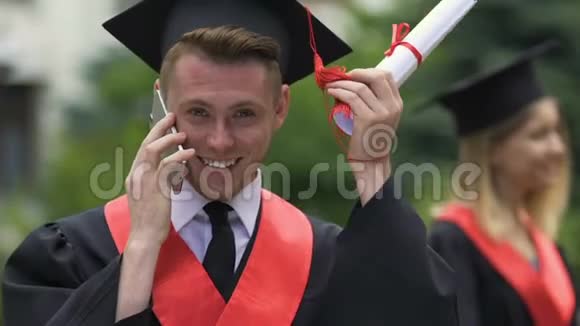 微笑的男毕业生拿着大学文凭在电话里交谈视频的预览图