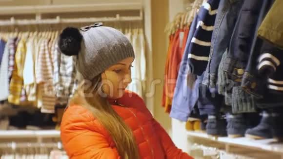 宝贝服装店里漂亮的女人穿着灰色针织帽子和红色夹克的白人女性看着婴儿衣服视频的预览图