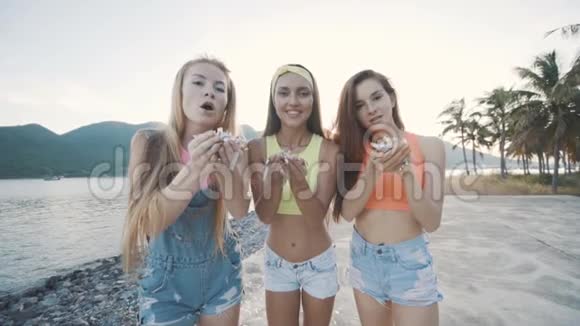 三个朋友的女人微笑着在日落时在海滩上吹着五颜六色的闪光视频的预览图