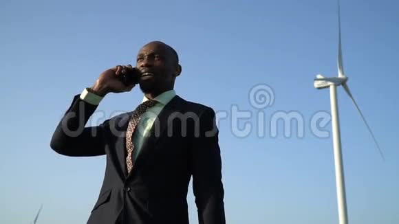 一个穿着商务服的黑人在风力发电机附近通过电话交流视频的预览图