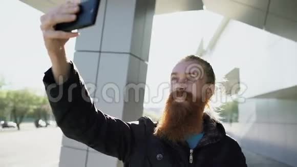 年轻的留胡子的嬉皮士用智能手机在户外拍摄自拍视频的预览图