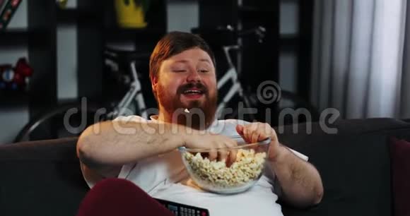 有胡子的胖子笑着在房间里看电视在桌子上吃爆米花和啤酒胖子喝啤酒概念视频的预览图