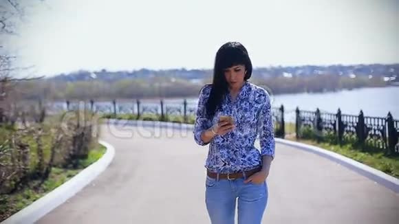 美丽的亚洲女孩穿着牛仔裤在阳光明媚的一天在公园散步在智能手机上阅读一条信息视频的预览图
