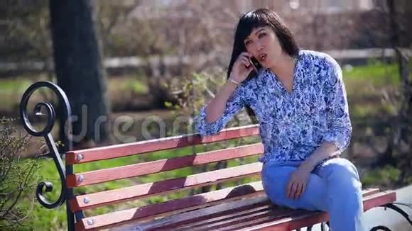 美丽的亚洲女孩坐在公园里坐在一张穿着蓝色牛仔裤的长凳上打电话视频的预览图