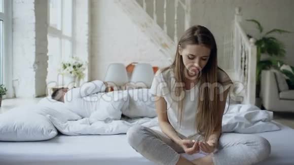 沮丧的年轻女子坐在床上哭泣而她的男朋友躺在家里的床上视频的预览图