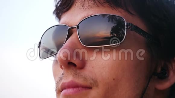 年轻英俊的时尚男士戴着太阳镜看起来很棒日落时带着耳机在热带海滩上听音乐享受着乐趣视频的预览图
