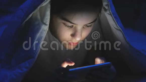 女孩晚上躺在毯子下在智能手机上聊天的特写镜头视频的预览图