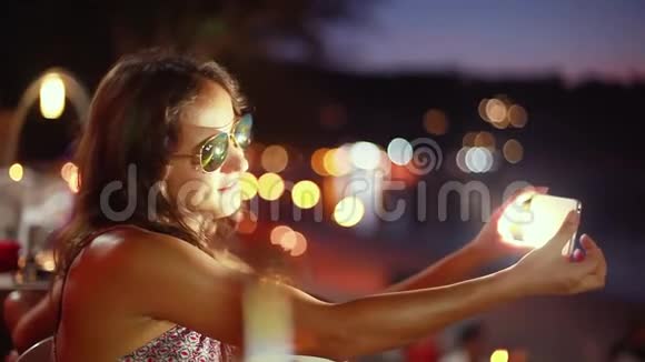 年轻漂亮的黑发女人戴着太阳镜摆着假脸用智能手机和闪光灯自拍视频的预览图