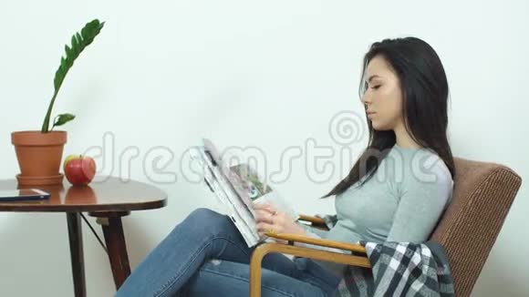 漂亮的女孩坐在舒适的椅子上在家看一本光滑的杂志视频的预览图