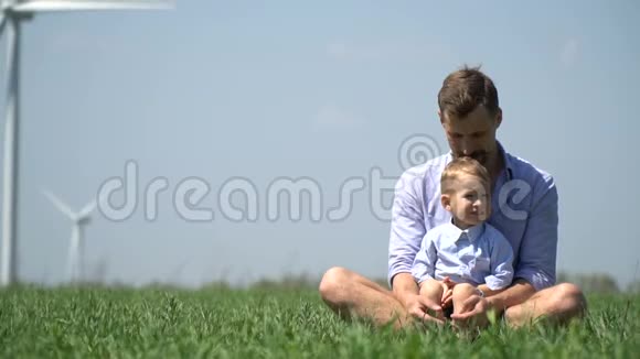 男孩坐在爸爸的身边在一片绿色草地的中间在一个风力发电机的背景下在一个视频的预览图
