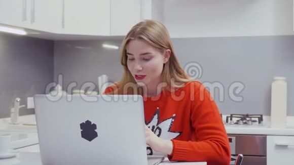 穿红色毛衣的漂亮白种人女孩在厨房里用笔记本电脑视频的预览图