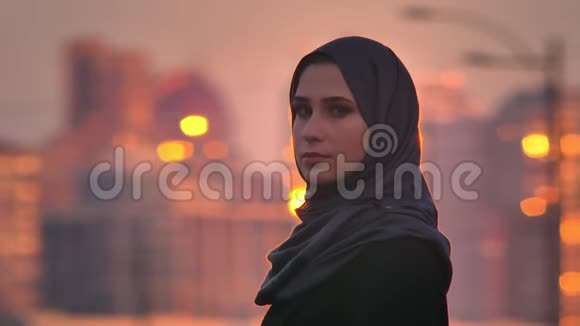 年轻迷人的穆斯林妇女戴着头巾在夕阳下看着美丽的城市视频的预览图
