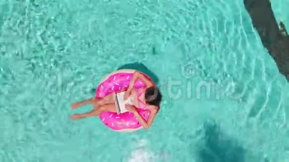 鸟瞰一个年轻的黑发女人游泳在一个充气大甜甜圈与笔记本电脑在一个透明的绿松石游泳池视频的预览图