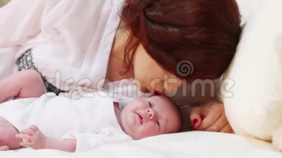 年轻的妈妈在卧室的白床上亲吻她可爱的小宝宝视频的预览图