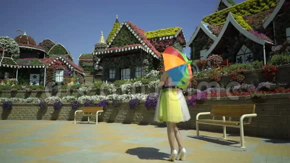 美丽的女孩穿着滑稽的复古连衣裙带着彩色彩虹伞在花园里散步女人正沿着视频的预览图