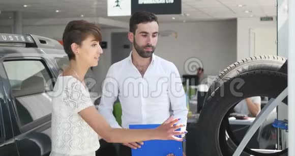 汽车服务公司一位年轻的汽车经理站在轮胎旁边和一位客户妇女讨论一些事情她和一位视频的预览图
