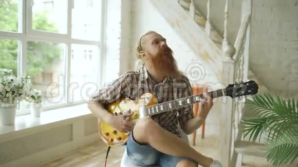 漂亮的胡须男坐在椅子上在家里的现代卧室里学弹吉他视频的预览图