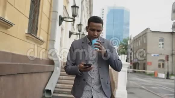 stadicam拍摄的年轻快乐商人使用智能手机在户外端着咖啡散步视频的预览图