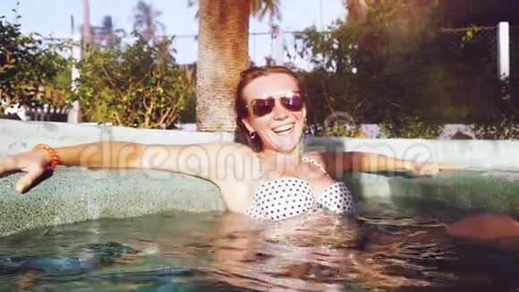 年轻漂亮的快乐女人戴着太阳镜在按摩浴缸里放松日落时分在室外游泳池享受她的生活视频的预览图