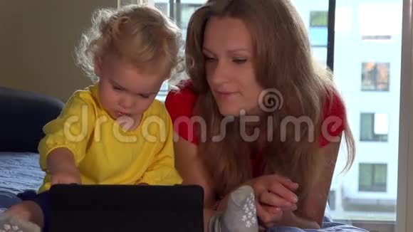 可爱的蹒跚学步的女孩和她的妈妈用平板电脑躺在沙发上微笑视频的预览图