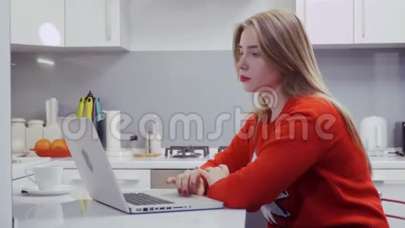 穿红色毛衣的漂亮女孩在厨房用笔记本电脑视频的预览图