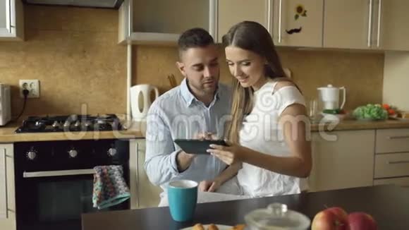 快乐的年轻夫妇在厨房里坐着早上吃早餐时使用数码平板电脑视频的预览图