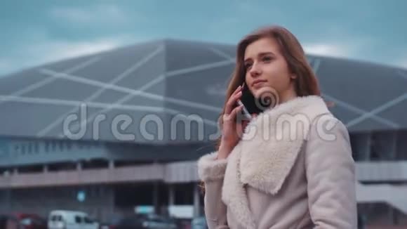 一个年轻漂亮的女孩站在一座现代化的大建筑前和一个伴侣在电话里交谈时的肖像视频的预览图