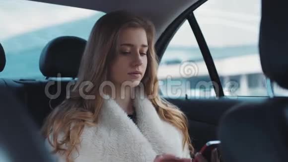 年轻漂亮的金发女郎开车在她的智能手机上打字与她的朋友交流视频的预览图