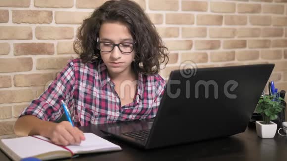 一个年轻漂亮的男孩头发卷曲黑发在笔记本电脑上工作在桌旁写字视频的预览图