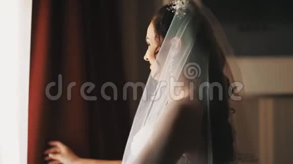 穿着婚纱的年轻新娘在半暗的房间里拉开窗帘视频的预览图