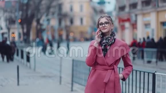 漂亮的年轻女子穿着一件优雅的粉红色外套站在市中心高兴地在电话里交谈拥挤的街道视频的预览图